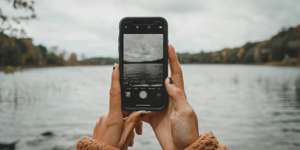 How the Pressure to Post on Social media is ruining travel and steps to overcome it - a person's hands holding a phone to click a picture of the lake